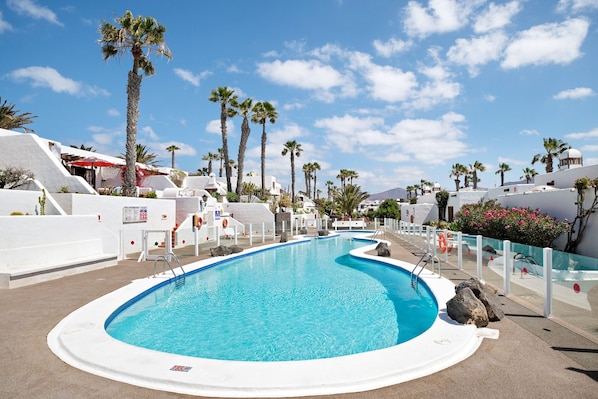 Une piscine entourée d’une terrasse blanche, de palmiers et de bâtiments blancs coiffés de toits en forme de dôme.