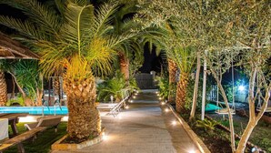 A nighttime outdoor pathway with palm trees, a pool, and benches. - Villa 'Tore' with Sea View, Wi-Fi, and Air Conditioning (Quartu Sant'Elena)