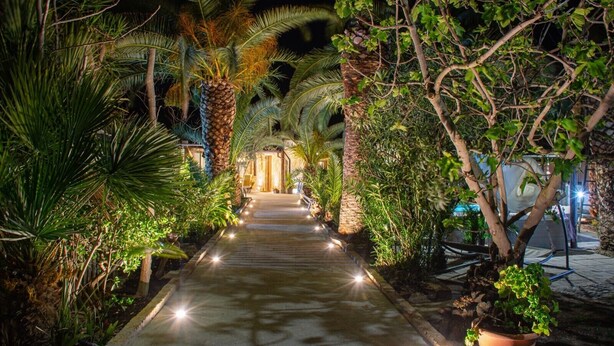 A nighttime pathway flanked by lush greenery and palm trees, leading to a building entrance. - Villa 'Tore' with Sea View, Wi-Fi, and Air Conditioning (Quartu Sant'Elena)