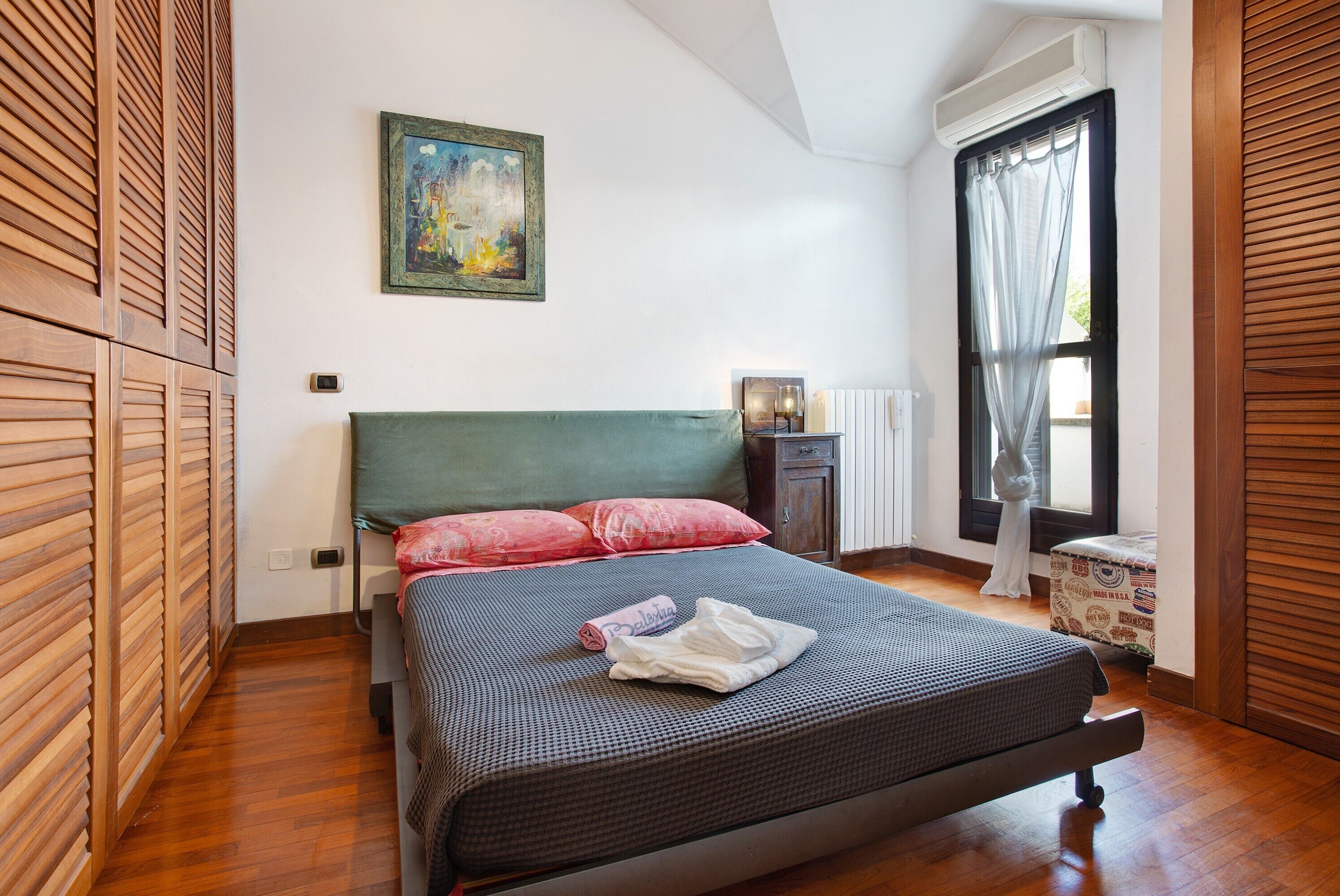 A bedroom with a bed, a painting on the wall, a window with curtains, and a radiator.
