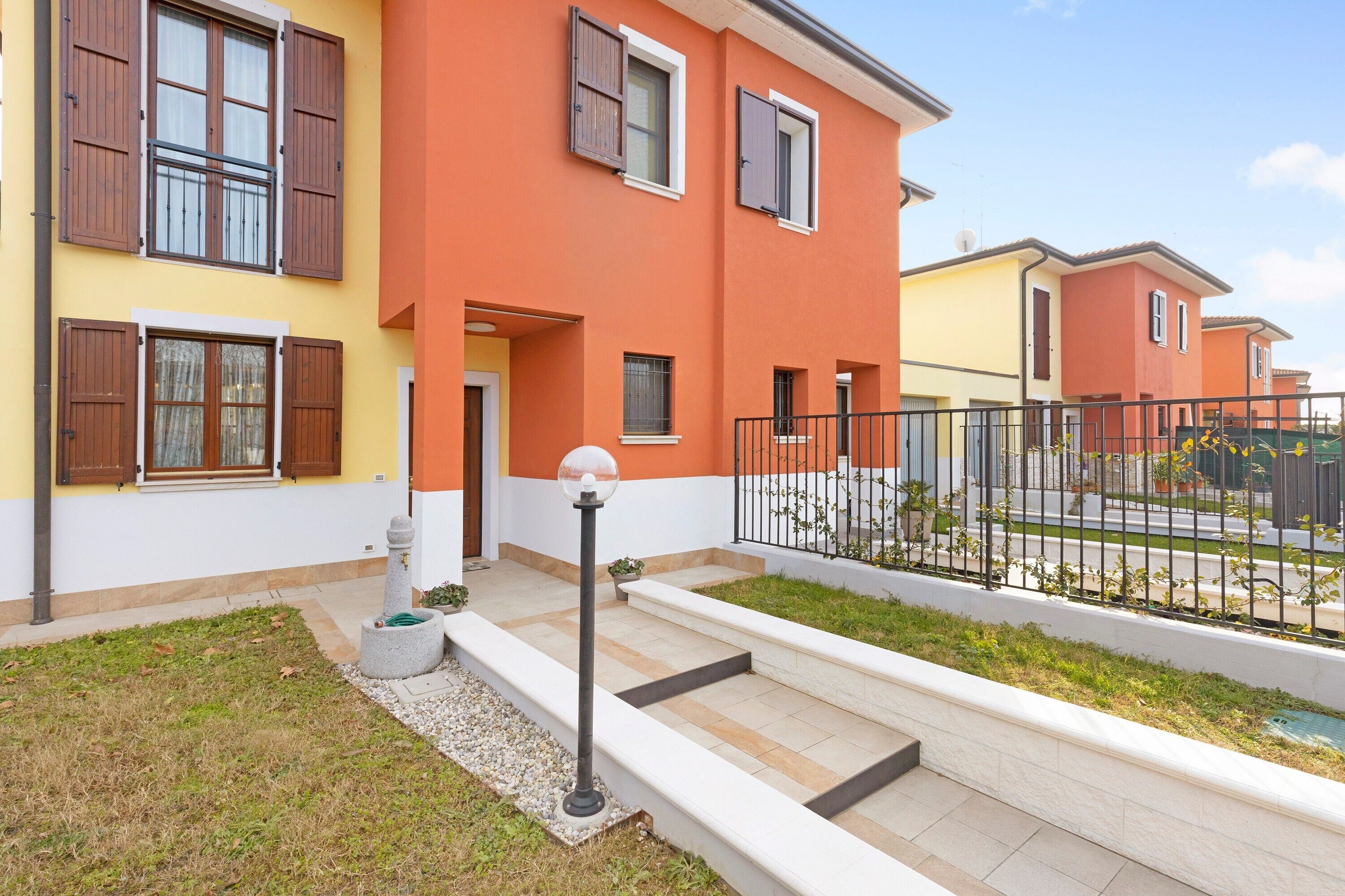 A residential building with a yellow facade, multiple windows with shutters, a visible entrance, and a small garden area with steps leading to the entrance.