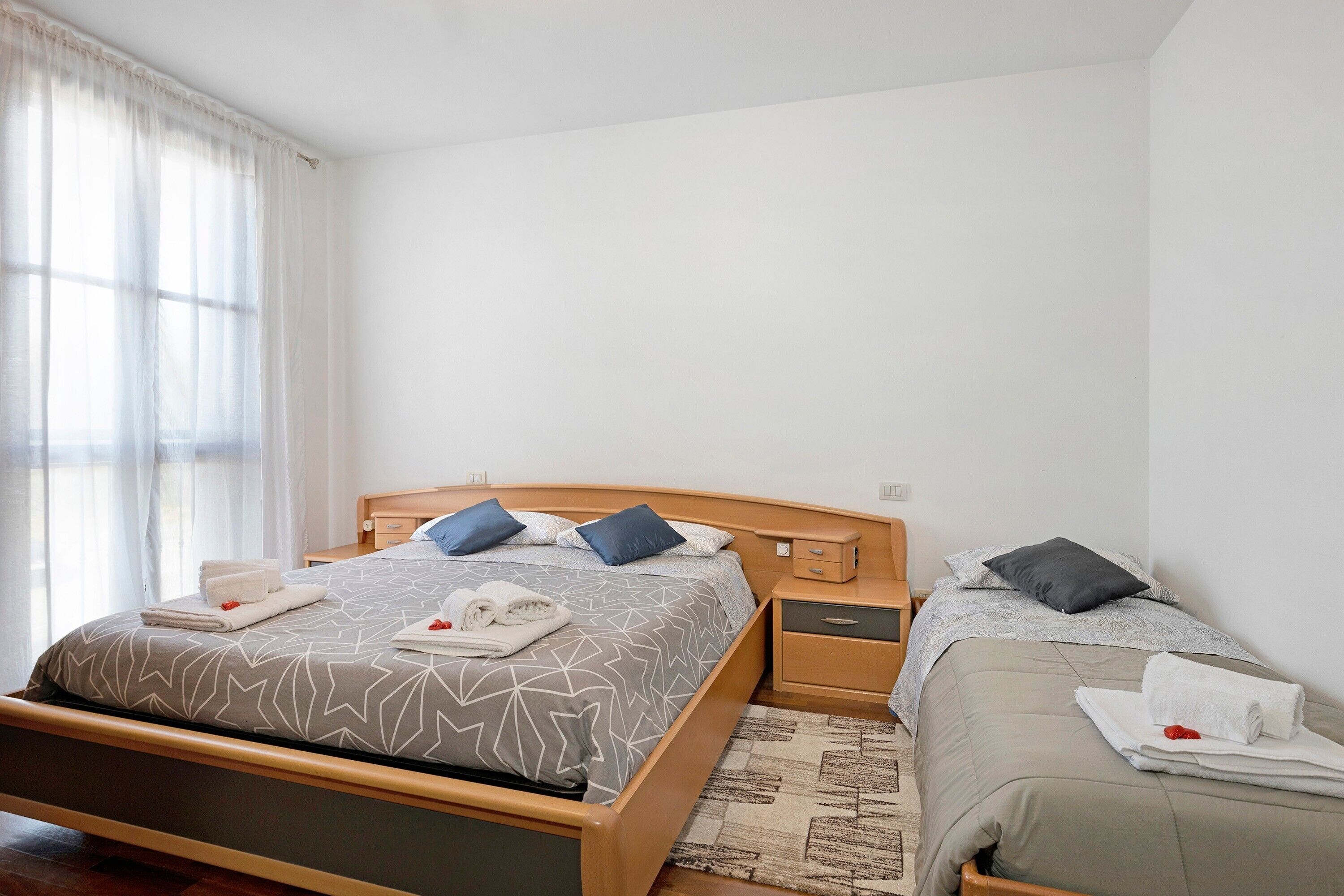 A double bed with a geometric patterned bedspread, two bedside tables, a wooden headboard, and a window with sheer curtains.