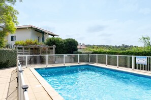 A swimming pool with a white fence, a house with a tiled roof, and a sign indicating a poolside café.