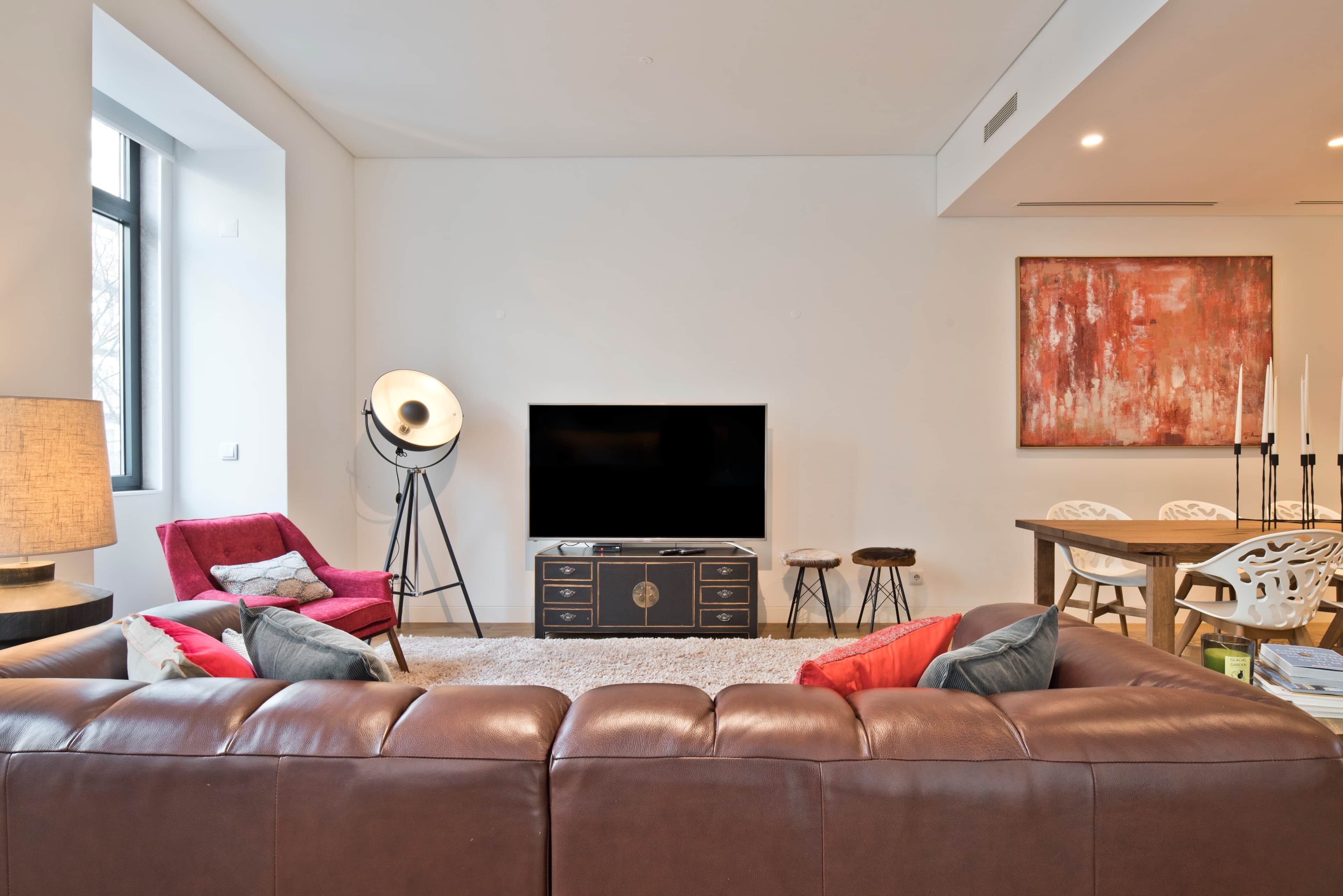 Un salon moderne avec un canapé en cuir, un fauteuil rouge, une lampe sur pied, un téléviseur, une table à manger avec des chaises et un grand tableau abstrait.