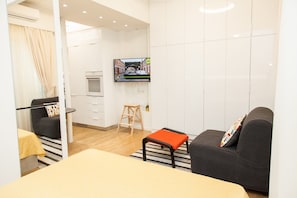 A modern hotel room with a bed, a sofa, a small stool, a TV mounted on the wall, and a kitchen area.