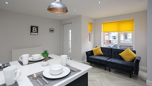 Een moderne keuken met een eethoek, een bank met gele kussens en uitzicht op de straat door grote ramen.