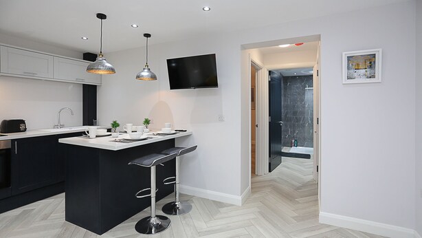 Een moderne keuken met een centraal eiland, zwarte barkrukken, hanglampen en een flatscreen televisie aan de muur.