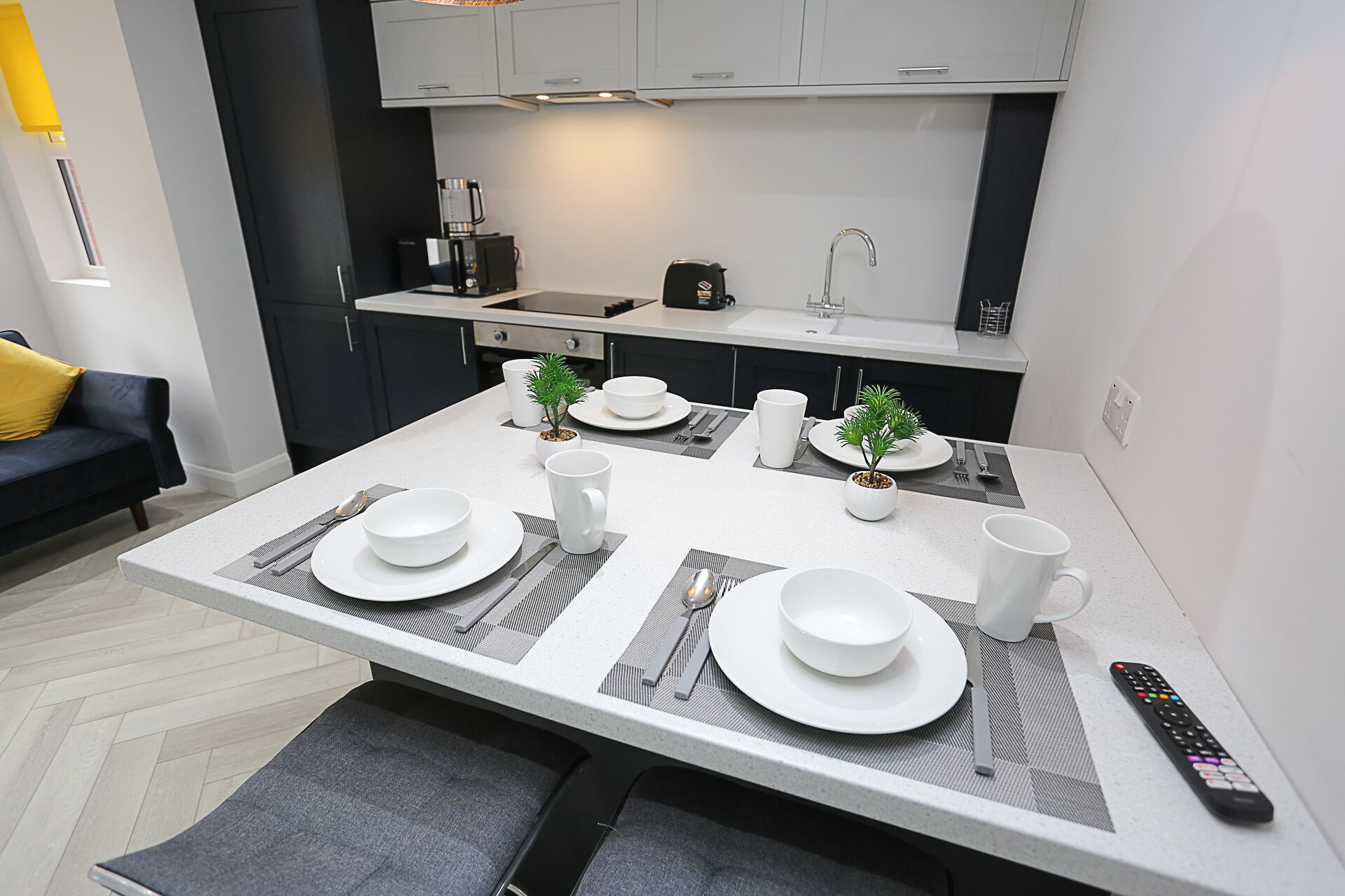Een moderne keuken met witte aanrechtbladen, een centraal kookeiland met eettafel, en een zwarte bank met een geel kussen op de achtergrond.