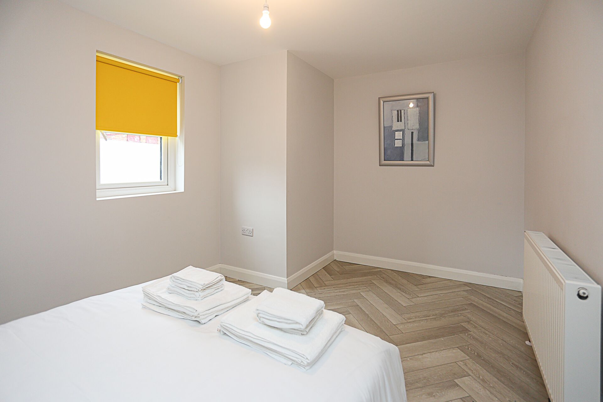 Ein Schlafzimmer mit einem Bett, einem Fenster mit gelbem Rollo, einer Heizung und einem gerahmten Bild an der Wand.