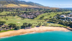 Vista aérea de un complejo turístico costero con campo de golf, playa y montañas al fondo.
