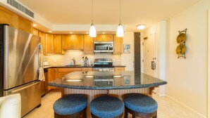 Cocina con gabinetes de madera, electrodomésticos de acero inoxidable, encimera de granito y tres taburetes de bar.