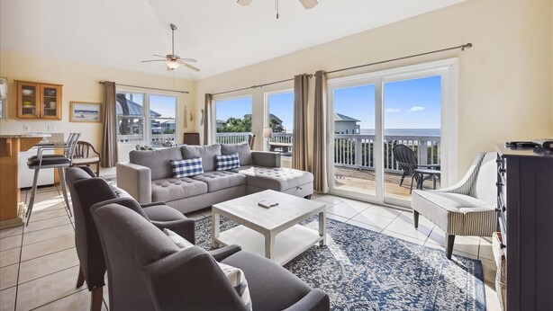 A spacious living room with a large sofa, a coffee table, and a ceiling fan. There is a sliding glass door leading to an outdoor patio area with a view of the ocean.