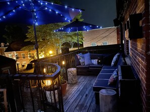 Una terraza de madera con mobiliario exterior, luces colgantes y un muro de ladrillo.