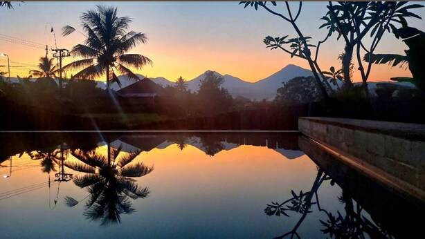 Una vista serena del atardecer sobre una masa de agua tranquila, con palmeras y montañas al fondo.