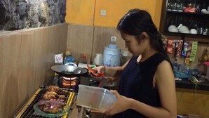 Una persona cocinando en una parrilla con una bandeja de comida. Se observa un dispensador de agua y una estantería con utensilios de cocina al fondo.