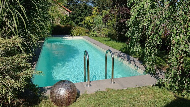 Une piscine rectangulaire avec des échelles en métal, entourée d’une végétation luxuriante et agrémentée d’une sphère décorative en pierre.