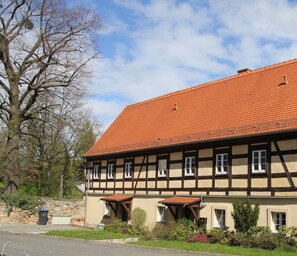 A traditional half-timbered building with a red-tiled roof, multiple windows, and a small garden area with various plants.