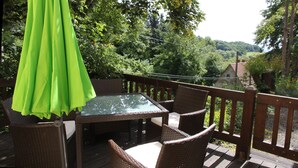 Une terrasse dotée de meubles en osier et d’une table en verre, entourée d’une végétation luxuriante et offrant une vue sur un village.