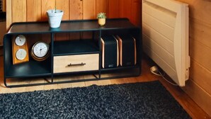 Een compacte kamer met houten paneelwanden, een radiator, een zwarte entertainmentkast met klokken en een kleine plant, en een donker vloerkleed.