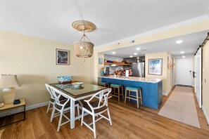 A dining area with a glass table and white chairs, a kitchen with a blue island, and a hallway leading to a door.