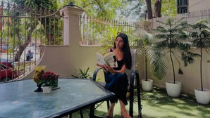 A person seated at a table with a book, surrounded by potted plants and a metal fence.