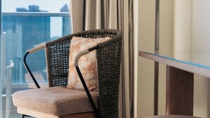 Une chaise en osier avec un coussin, une table en verre, une lampe et une vue sur la ville par une fenêtre.
