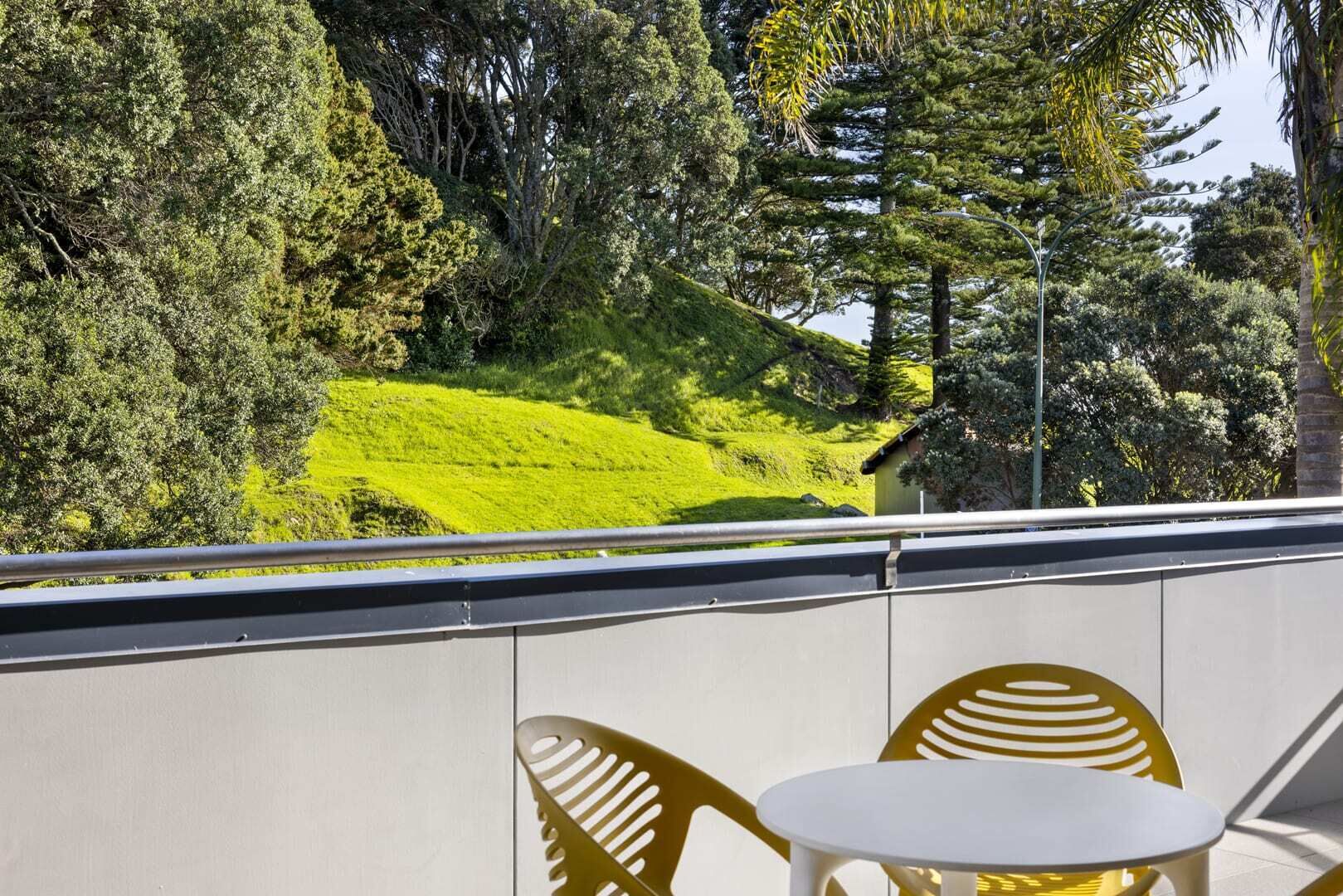 Ein Balkon mit Blick auf einen grünen Hang und Bäume. Zwei gelbe Stühle und ein kleiner weißer Tisch sind vorhanden.