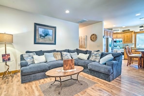 A living room with a sectional sofa, a round coffee table, a framed picture of an elk, and a kitchen area with wooden cabinets and a clock.