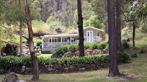 A small, single-story house with a white exterior and a balcony, surrounded by lush greenery and trees. - Cute Cottage with Log Burning Fire, close to Wineries (Megalong Valley)