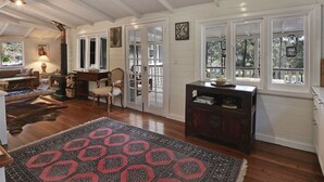 A spacious living room with a fireplace, wooden flooring, and a large rug. There are multiple windows and a view of the outdoors. - Cute Cottage with Log Burning Fire, close to Wineries (Megalong Valley)