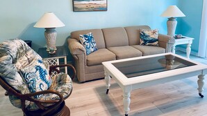 A living room with a beige sofa, a wooden rocking chair, a glass coffee table, two lamps, and a beach-themed painting on the wall.