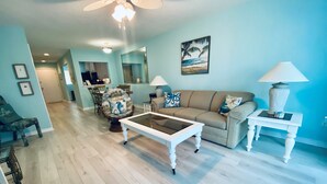A spacious living room with a light blue wall, a wooden floor, a white coffee table, a beige sofa, and a dining area in the background.
