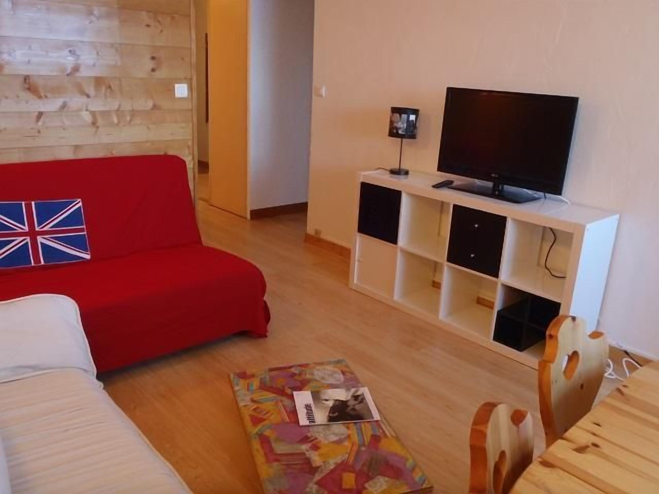 Un salon avec un canapé rouge, un canapé blanc, une table basse en bois et un téléviseur posé sur un meuble blanc.