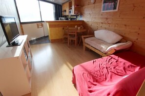 A compact room with a bed, a small table, a chair, a television, and a kitchenette.