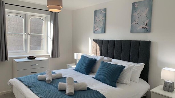 Un letto rifatto con lenzuola e cuscini blu, un comodino con una lampada e un quadro appeso al muro.