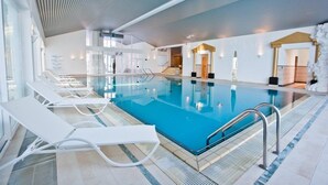 Ein überdachter Pool mit weißen Liegestühlen, klares blaues Wasser und modernes Architekturdesign mit schräg verlaufender Decke und Glaswänden.