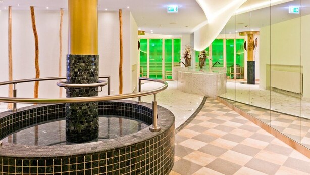 Eine moderne Lobby mit einem zentralen Wasserspiel, einem Schachbrettmuster-Boden und einem im Hintergrund grün beleuchteten Raum.