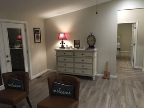 A room with a wooden floor, a white dresser with drawers, a lamp, a clock, and two chairs with 'welcome' pillows.