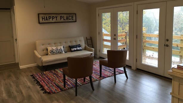 Ein Wohnzimmer mit einem weißen Sofa, zwei braunen Sesseln, einem bunten Teppich, einem Deckenventilator und einem Schild mit der Aufschrift 'HAPPY PLACE'.