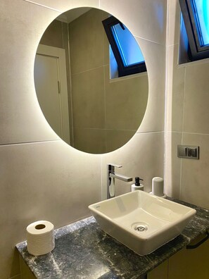 A bathroom with a round mirror, a white sink, and a toilet paper roll on the counter.