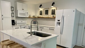 Uma cozinha moderna com bancada de mármore, pia, banquetas de madeira, geladeira branca e micro-ondas.