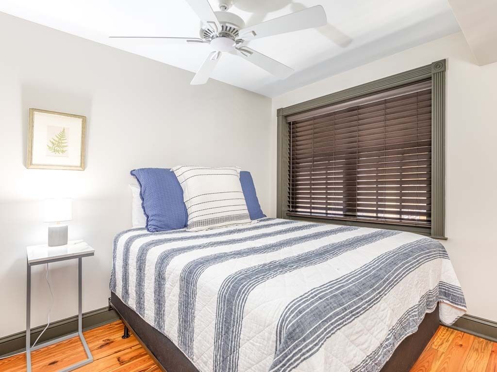 Un dormitorio con cama, ventilador de techo, mesita de noche con lámpara y ventana con persianas.