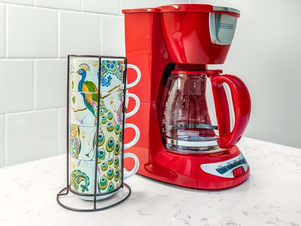 A red coffee maker and a colorful mug stand with a peacock design.