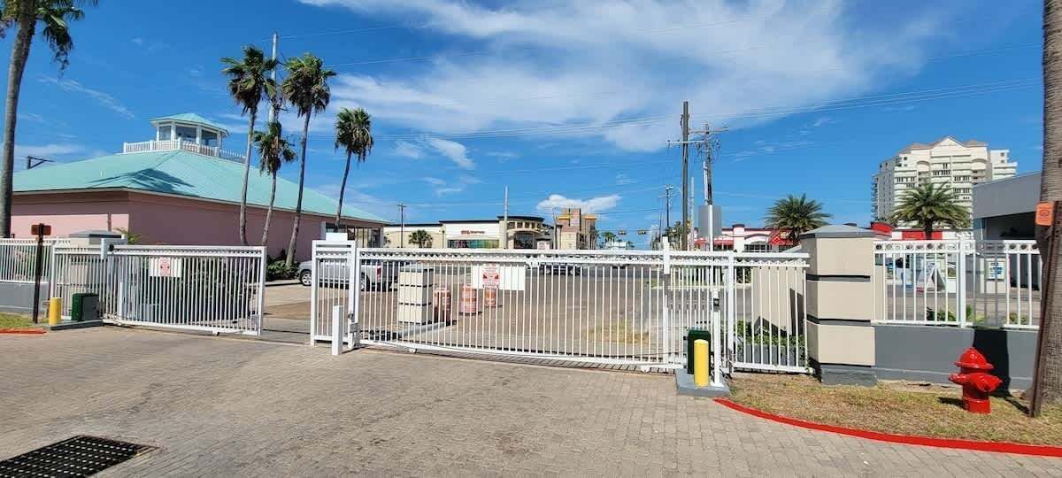 A gated entrance with a metal fence, palm trees, and buildings in the background.