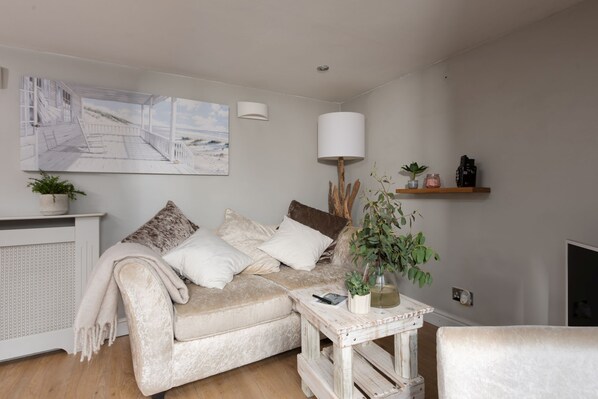 A cozy living room with a beige sofa, a small side table, a lamp, and a painting of a beach scene.