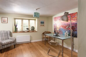 A cozy room with a plaid sofa, a table with a chair, a radiator, a window with blinds, and a colorful abstract painting. - A cosy cottage with a woodburner (Gwithian)