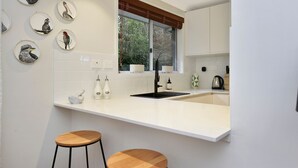 Eine moderne Küche mit weißer Arbeitsplatte, zwei Barhocker, einem Fenster mit Jalousien und an der Wand befestigten Tellern mit Vogelmotiven.
