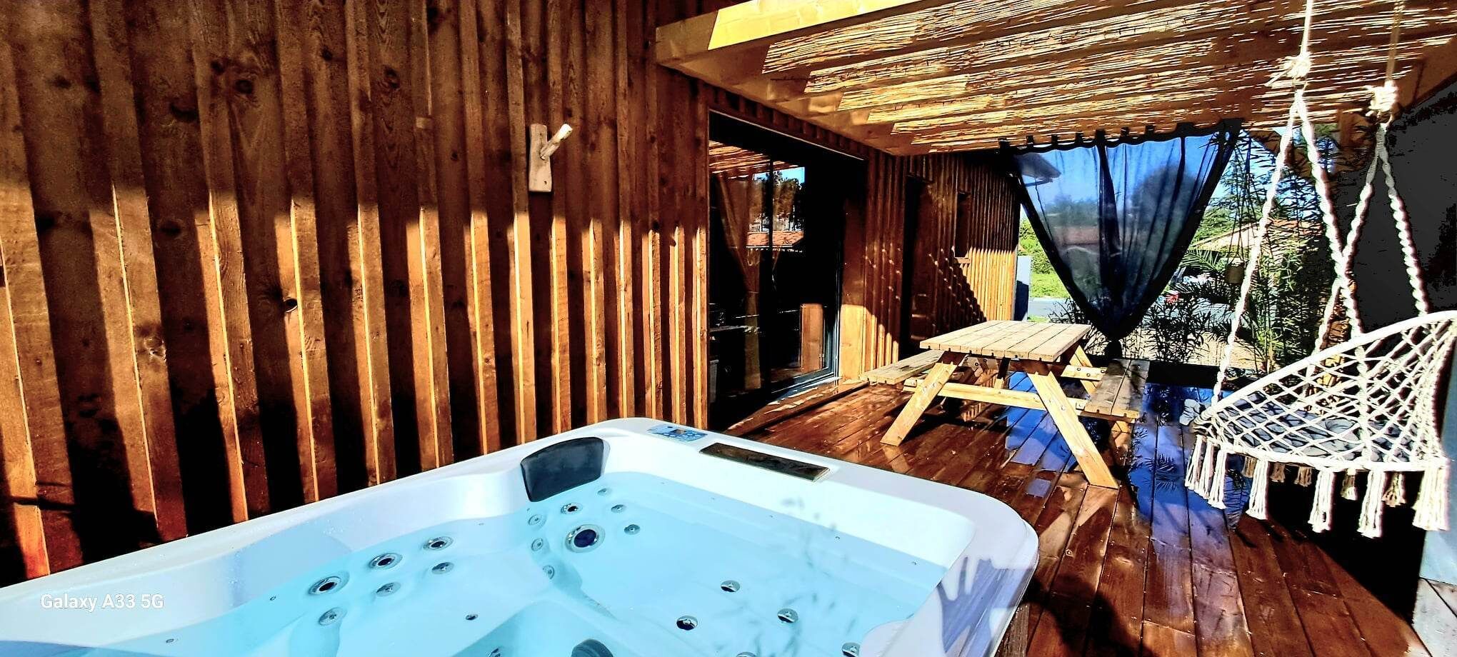 Une terrasse en bois avec un jacuzzi, une table en bois et un fauteuil suspendu.