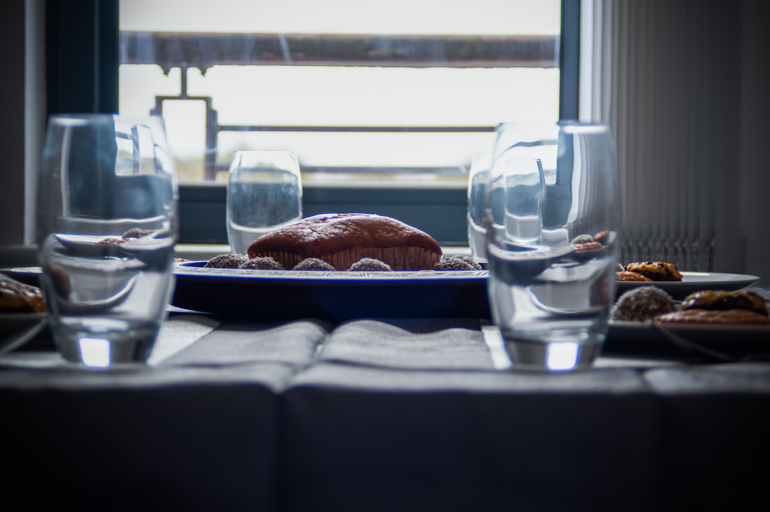 A table with a muffin on a plate, surrounded by clear glasses of water, set by a window with a view.