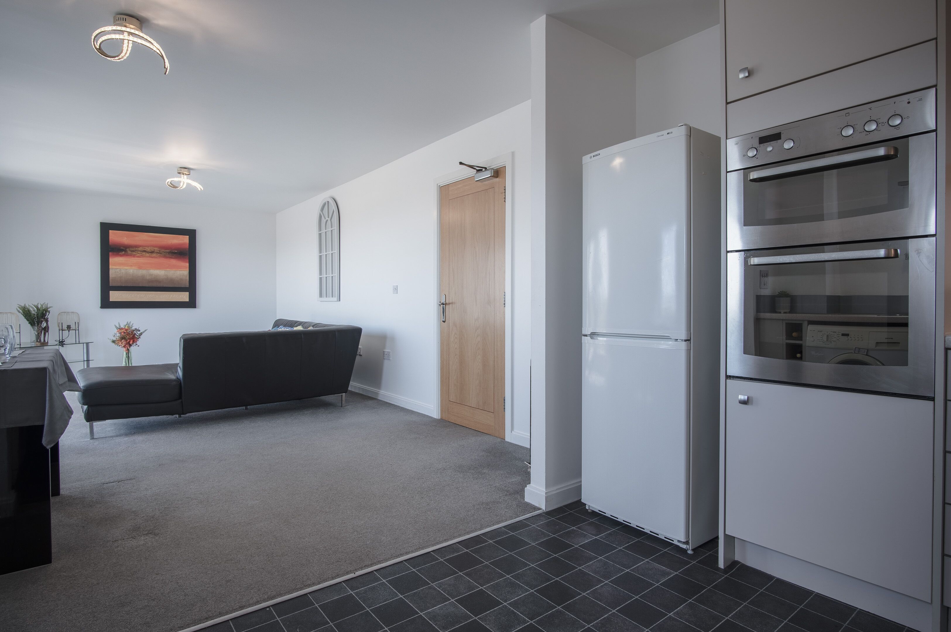 A modern kitchen with a built-in oven and refrigerator, a grey sofa, and a wooden door.